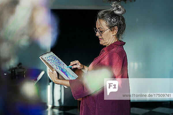 Ältere Frau mit Brille zeichnet in einem Buch  während sie zu Hause steht