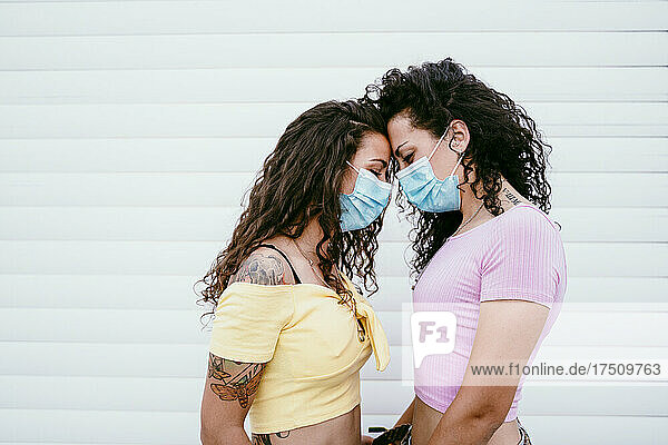 Lesbian couple wearing masks with face to face standing against wall in city