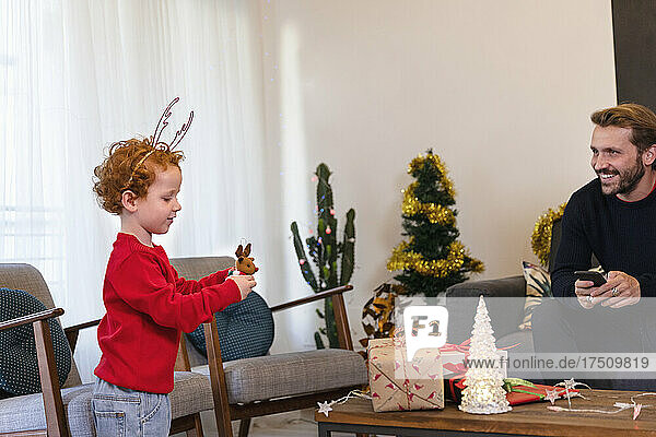 Smiling father looking at cute son wearing antler's headband playing with toy in living room