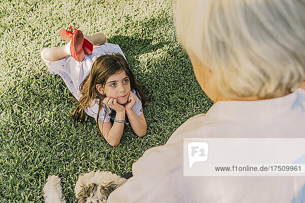 Cute girl with hands on chin looking at grandmother while lying over grassy land in yard