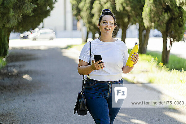Smiling curvy young woman with mobile phone and bottle in a public park