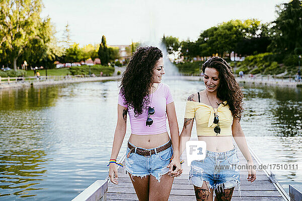 Lesbian couple holding hands while walking on pier against lake