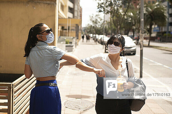 Freundinnen mit Sonnenbrille und Maske geben beim Stehen in der Stadt einen Ellbogenstoß