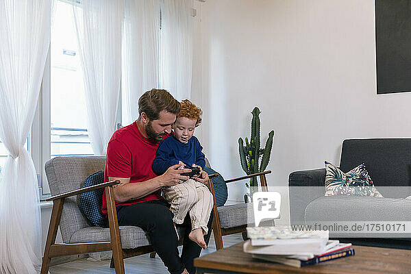Father with cute son holding joystick while playing video game in living room at home