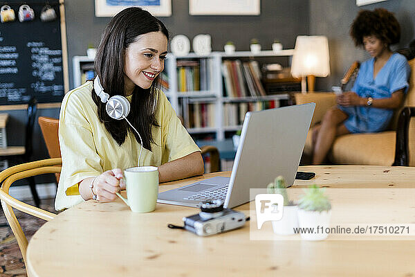 Happy woman using laptop at cafe with friend in background