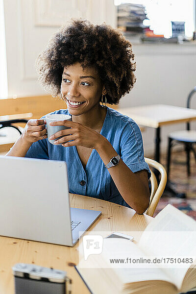 Thoughtful woman holding coffee mug while sitting with laptop at coffee shop