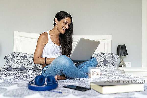 Smiling woman using laptop while sitting with book and headphones on bed at home