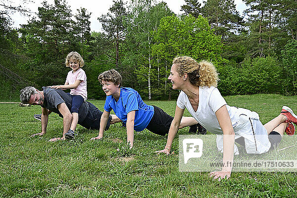 Girl sitting on father back exercising with family over grassy land in forest