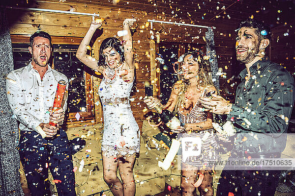Happy friends opening champagne while dancing amidst confetti in party