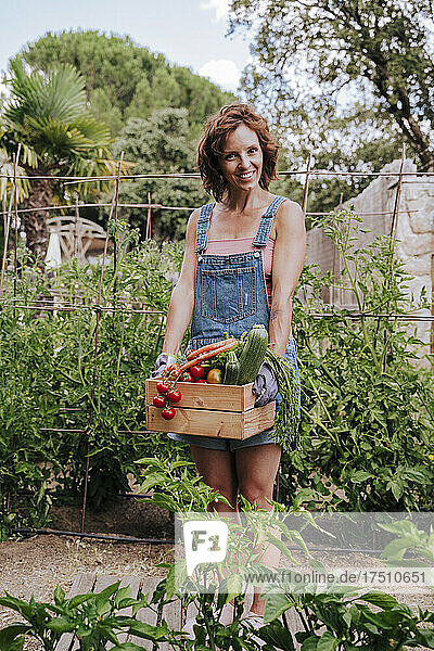 Smiling woman holding various vegetables in crate while standing against plants at community garden