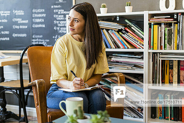 Thoughtful woman writing in book while sitting on chair in coffee shop