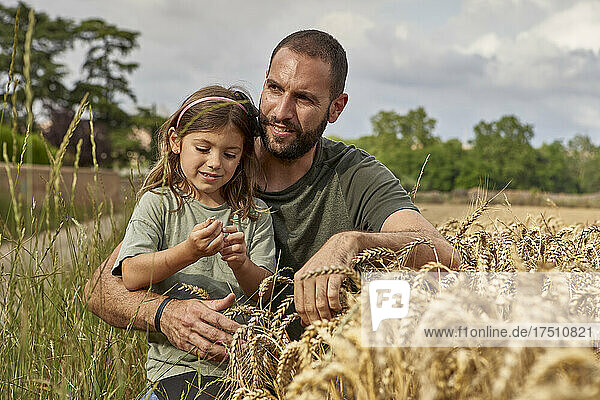 Father with daughter sitting by crops in wheat field against sky