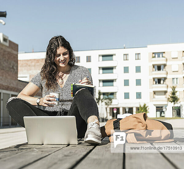 Smiling woman holding drink writing in book while sitting on boardwalk during sunny day