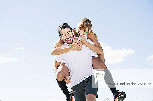 Cheerful man piggybacking woman while walking against sky during sunny day