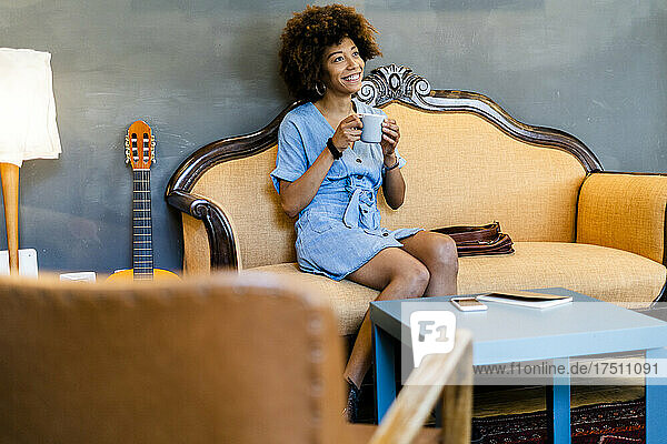 Thoughtful woman holding coffee mug while sitting on sofa in modern cafe