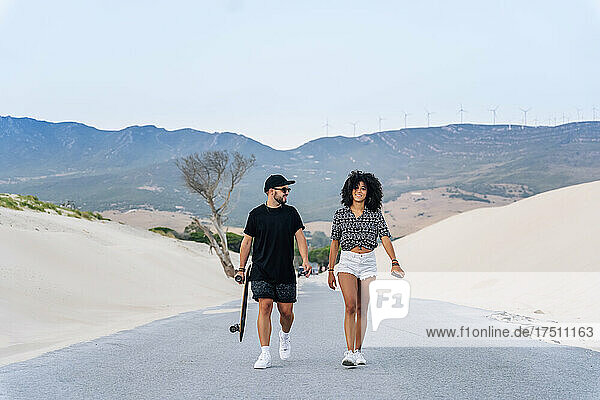 Smiling multi-ethnic couple walking on road amidst sand dunes against clear sky