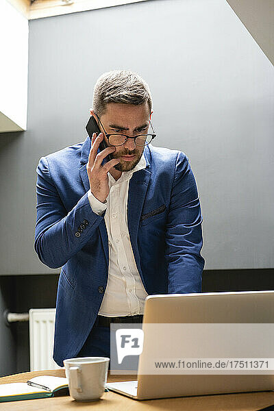 Businessman talking on smart phone while using laptop at desk in creative office