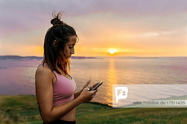 Fit young woman using smart phone while by sea during sunset