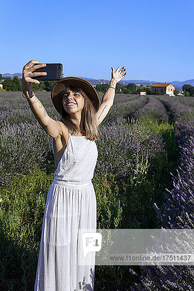 Smiling woman wearing white dress taking selfie with smart phone while standing amidst lavender field