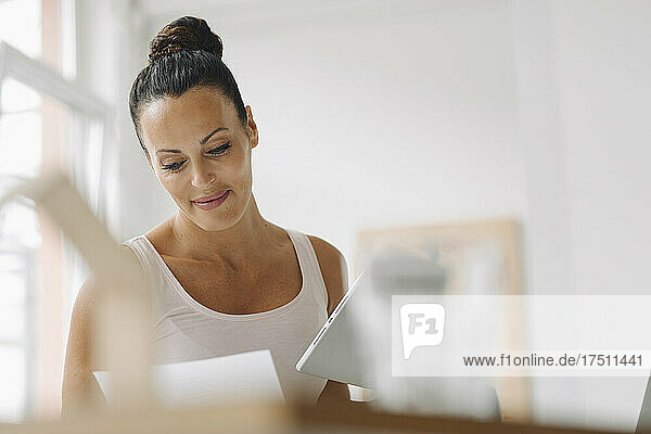 Smiling woman holding digital tablet reading document while sitting at home office