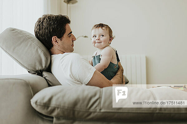 Father holding cute baby daughter while relaxing on sofa at home