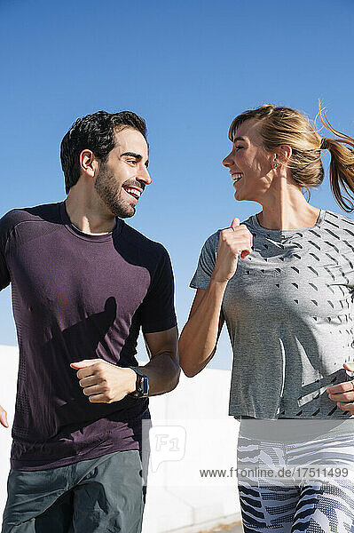 Smiling couple looking at each other while running against clear blue sky on sunny day