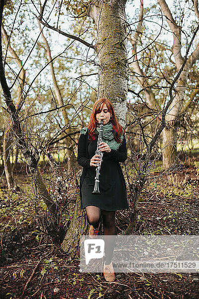 Beautiful young woman practicing clarinet while leaning on bare tree trunk in forest