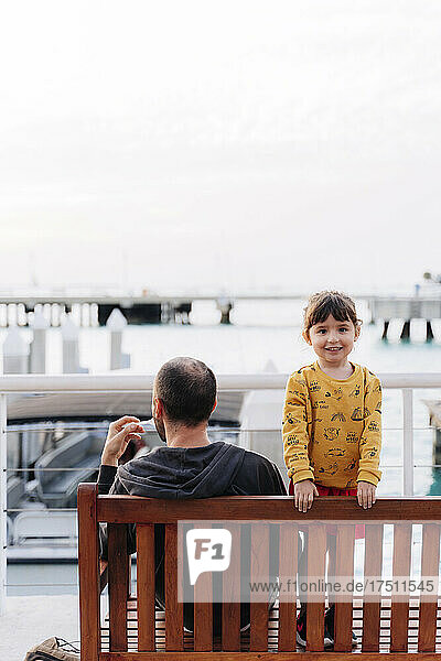 Smiling girl standing by father sitting on bench against sea