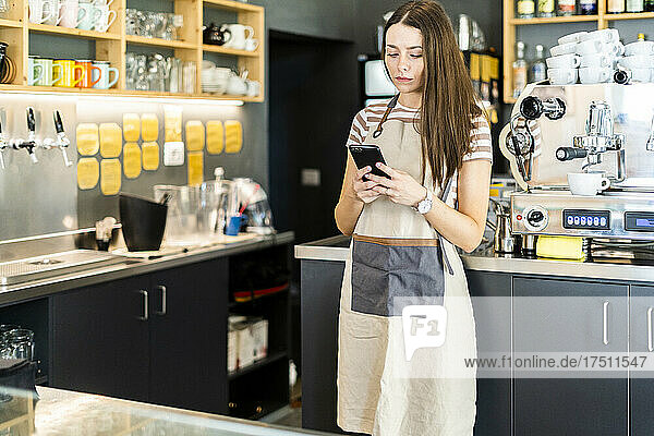 Young female owner using smart phone while standing in coffee shop