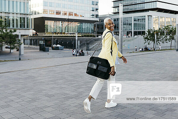 Smiling businesswoman with phone and bag walking in city