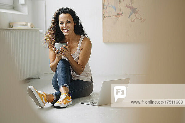 Smiling beautiful woman holding coffee mug while using laptop on floor at home