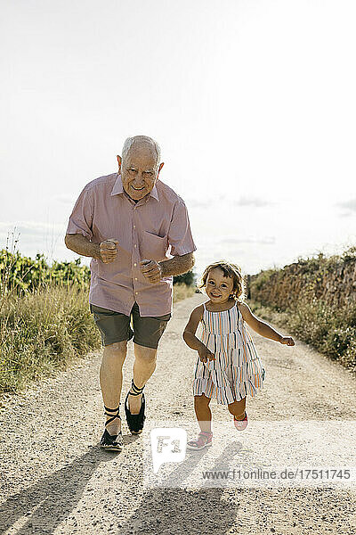 Playful senior man running with granddaughter on dirt road amidst plants during sunny day