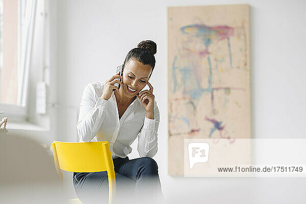 Lächelnde Geschäftsfrau spricht über Smartphone  während sie im Heimbüro auf einem Stuhl sitzt