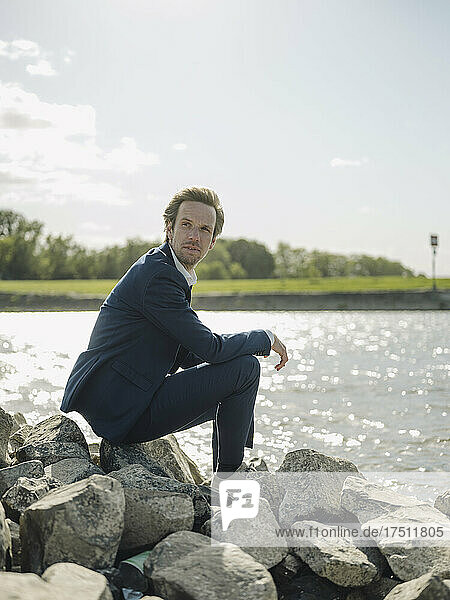 Thoughtful businessman sitting on rock at riverbank against sky during sunny day