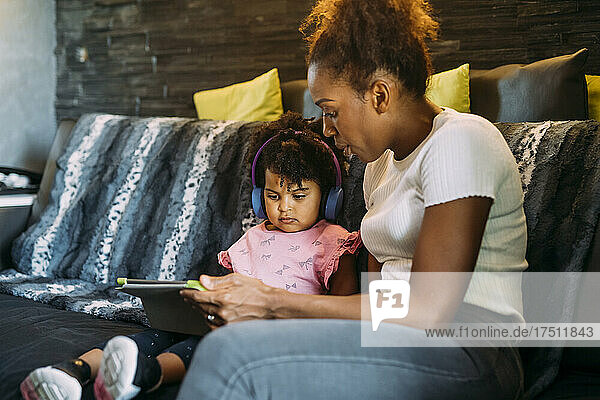 Mutter zeigt Tochter mit Kopfhörern digitales Tablet  während sie zu Hause auf dem Sofa sitzt