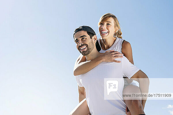 Mid adult man piggybacking cheerful woman against clear sky during sunny day