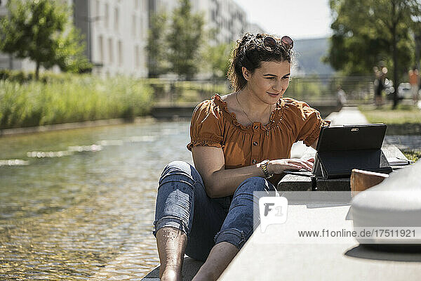 Woman using laptop while sitting by pond in park on sunny day