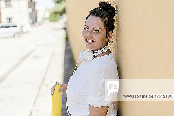 Portrait of a smiling curvy young woman with bottle and headphones leaning against a wall
