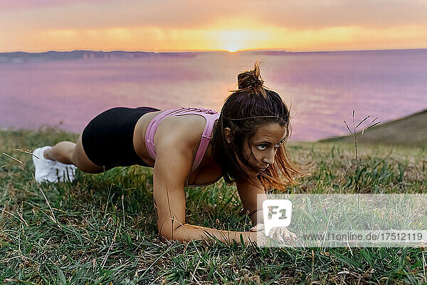 Determined young active woman practicing plank position on grass during sunset