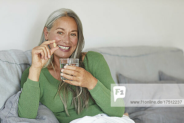 Smiling woman showing pill with drinking water while sitting on sofa at home