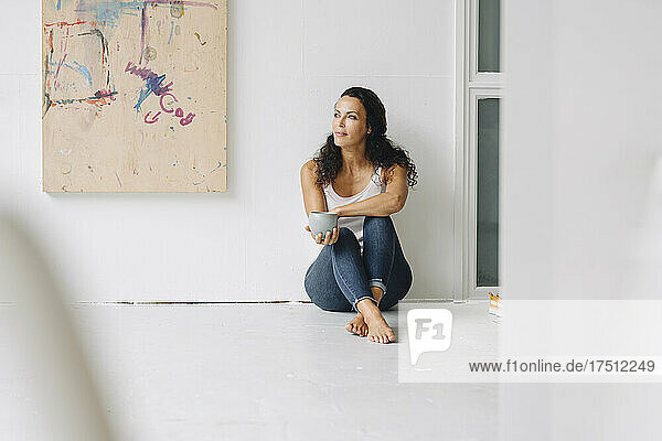 Frau hält Kaffeetasse in der Hand und denkt nach  während sie auf dem Boden an der Wand im Dachgeschoss sitzt
