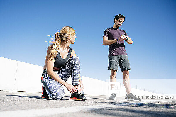 man checking time while woman tying shoelace on road against clear blue sky in city