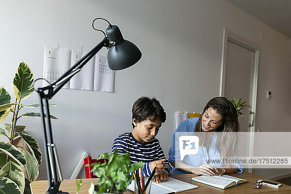 Smiling female tutor looking at boy studying on table