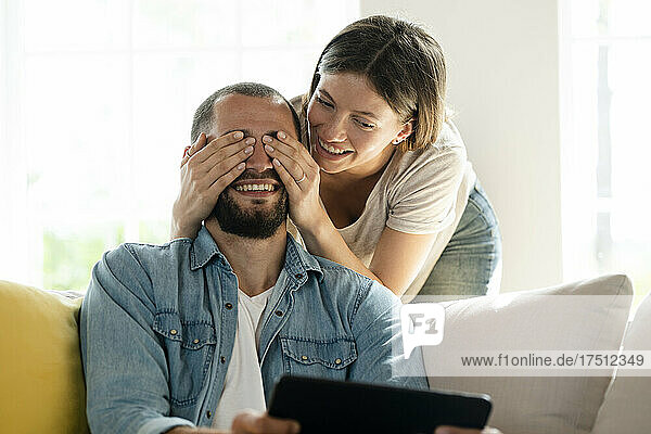 Young woman surprising man  covering his eyes  while he is sitting on the couch