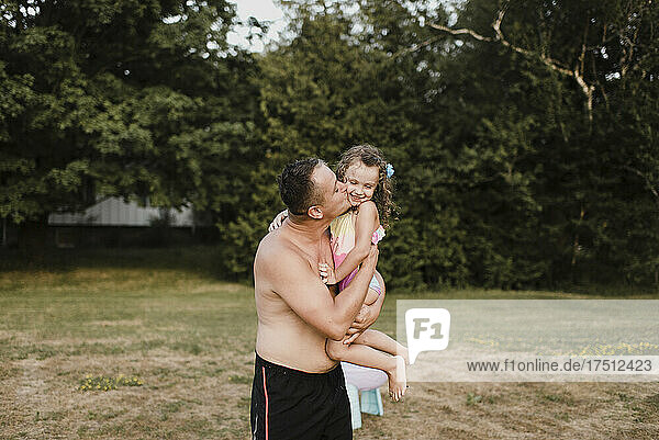 Father wearing swimming trunks carrying and kissing little daughter on a meadow