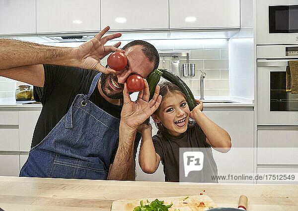 Cheerful father and daughter playing with tomatoes and zucchinis in kitchen