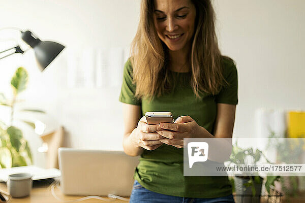 Smiling young woman using smart phone while standing at desk in home office