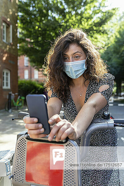 Junge Frau mit Maske macht ein Selfie mit dem Smartphone  während sie auf dem Fahrrad am Parkplatz sitzt