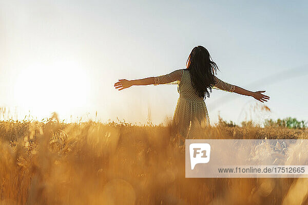 Young woman with arms outstretched standing amidst wheat farm against clear sky during sunset