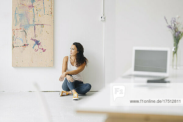 Thoughtful mid adult woman looking away while sitting on floor against wall at home
